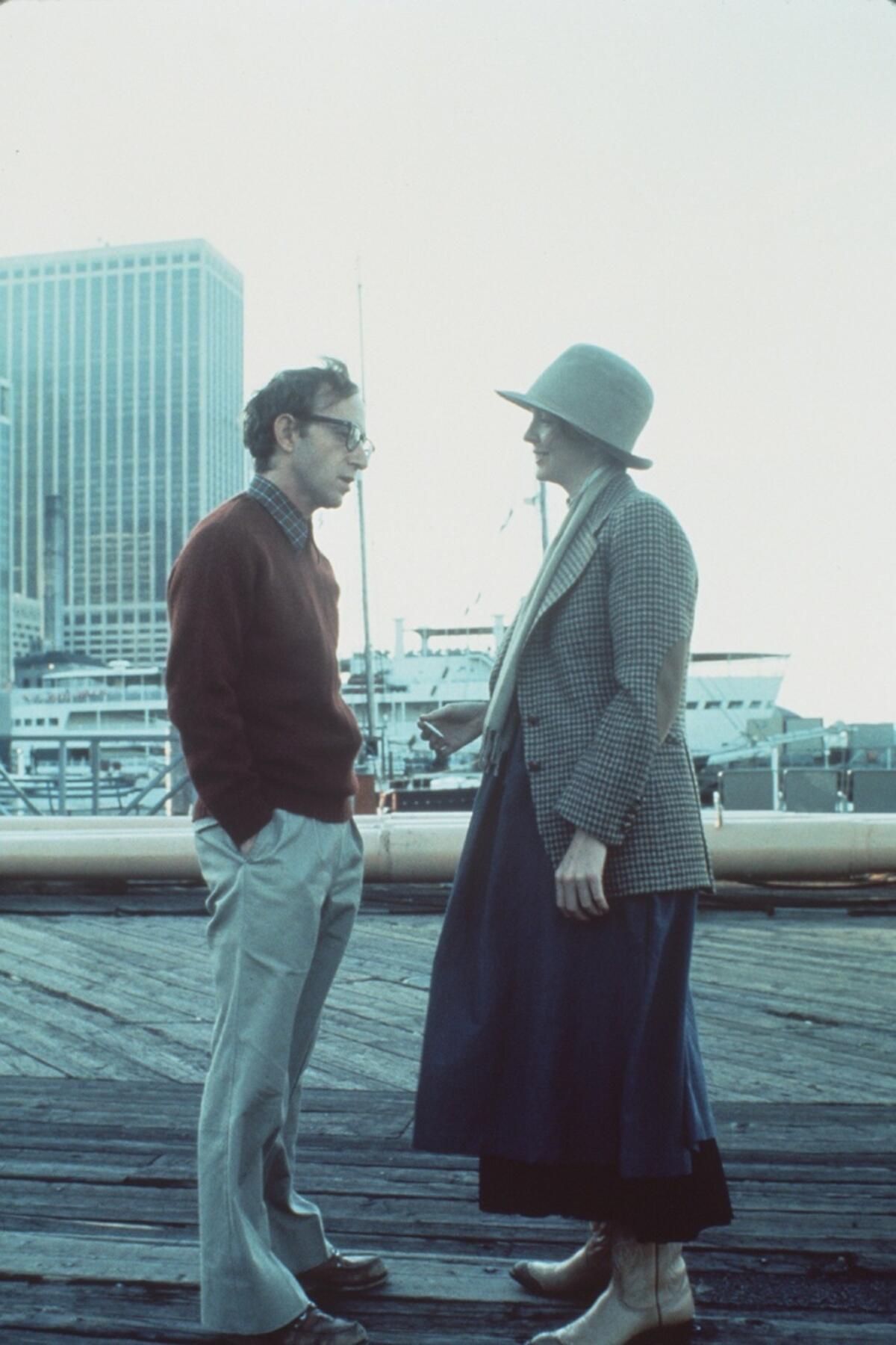 A man and woman talk outside with a tall building looming behind them.