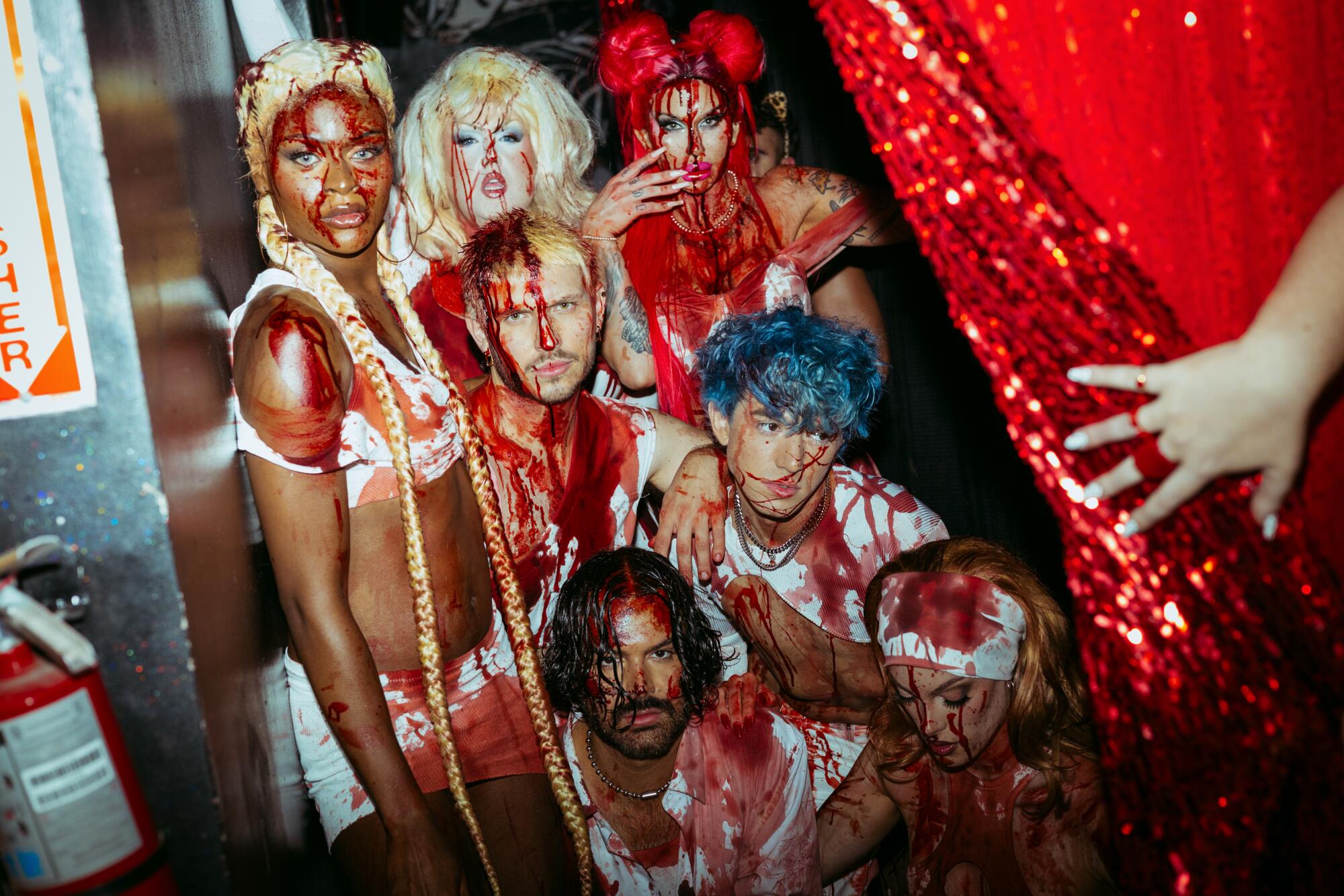A hand pulls back a spangly curtain to reveal people dressed in white and covered in fake blood.