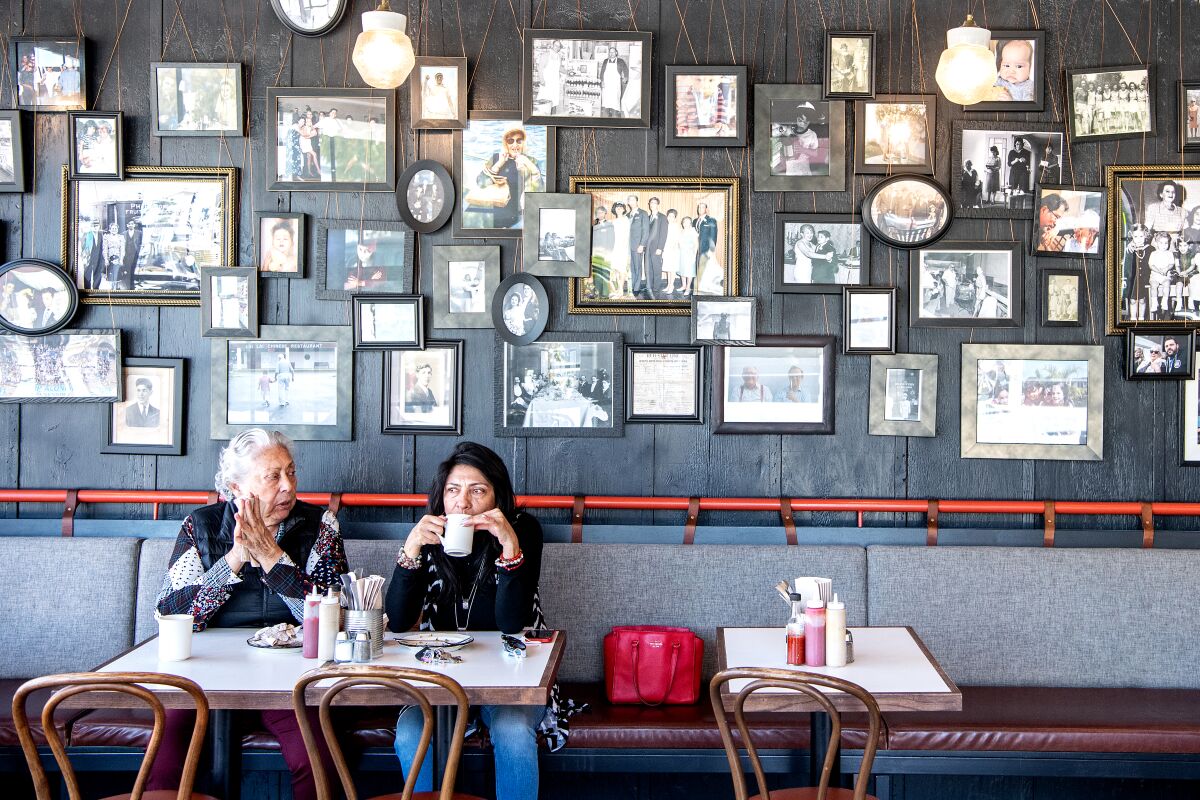 Two people sit in a restaurant against a wall displaying many family photos