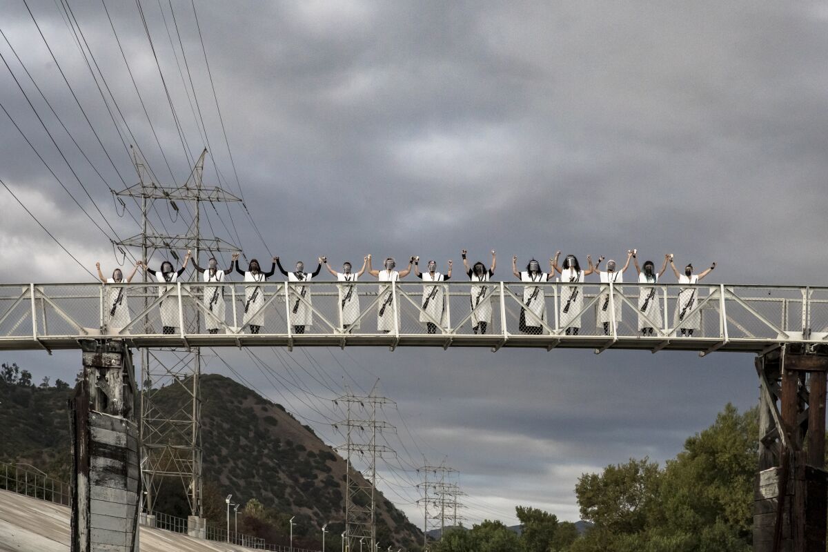 Women in white caftans emblazoned with an image of a rifle stand triumphantly on a pedestrian bridge over the L.A. River