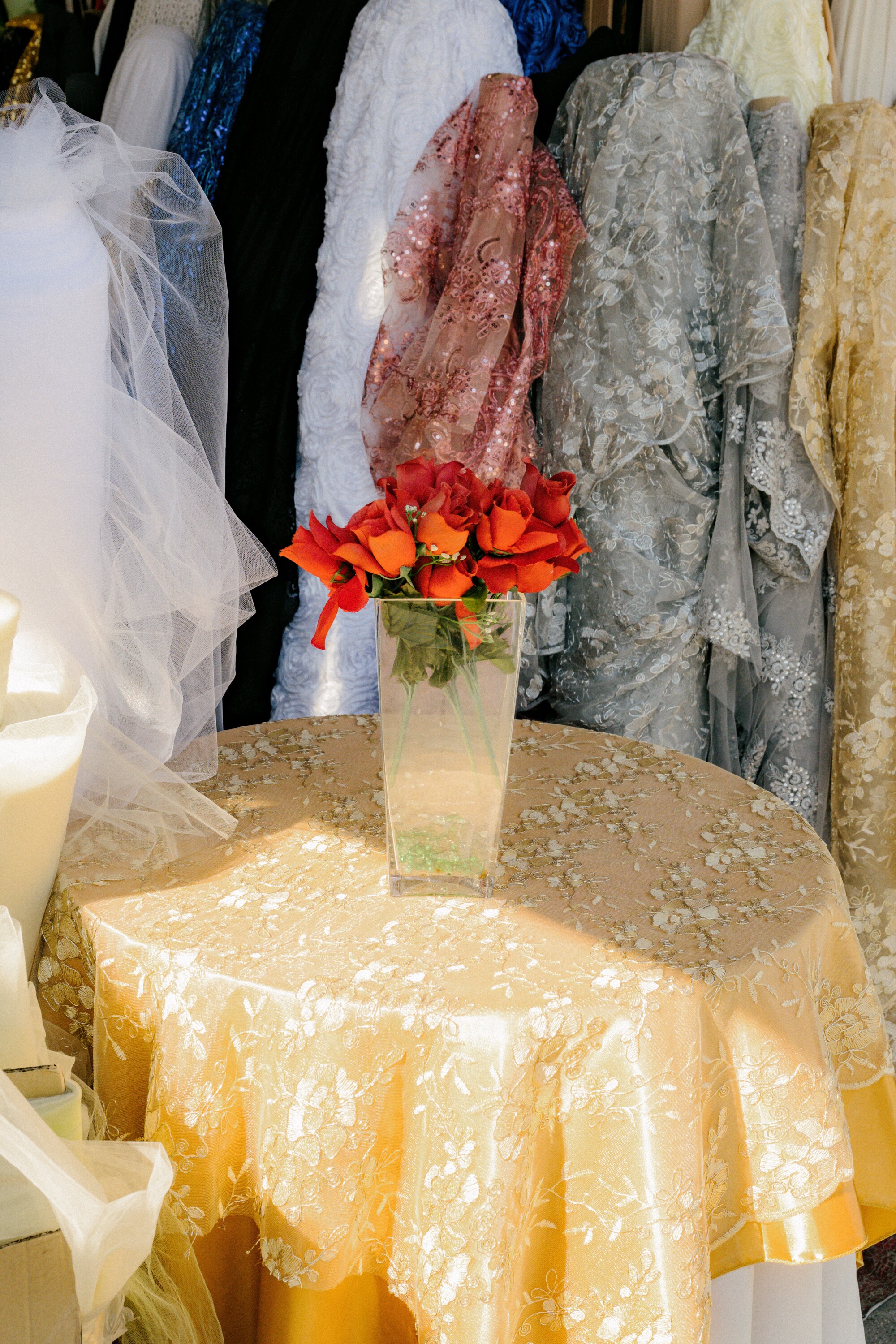 A vase of fake roses on a table with bolts of fabric behind it.