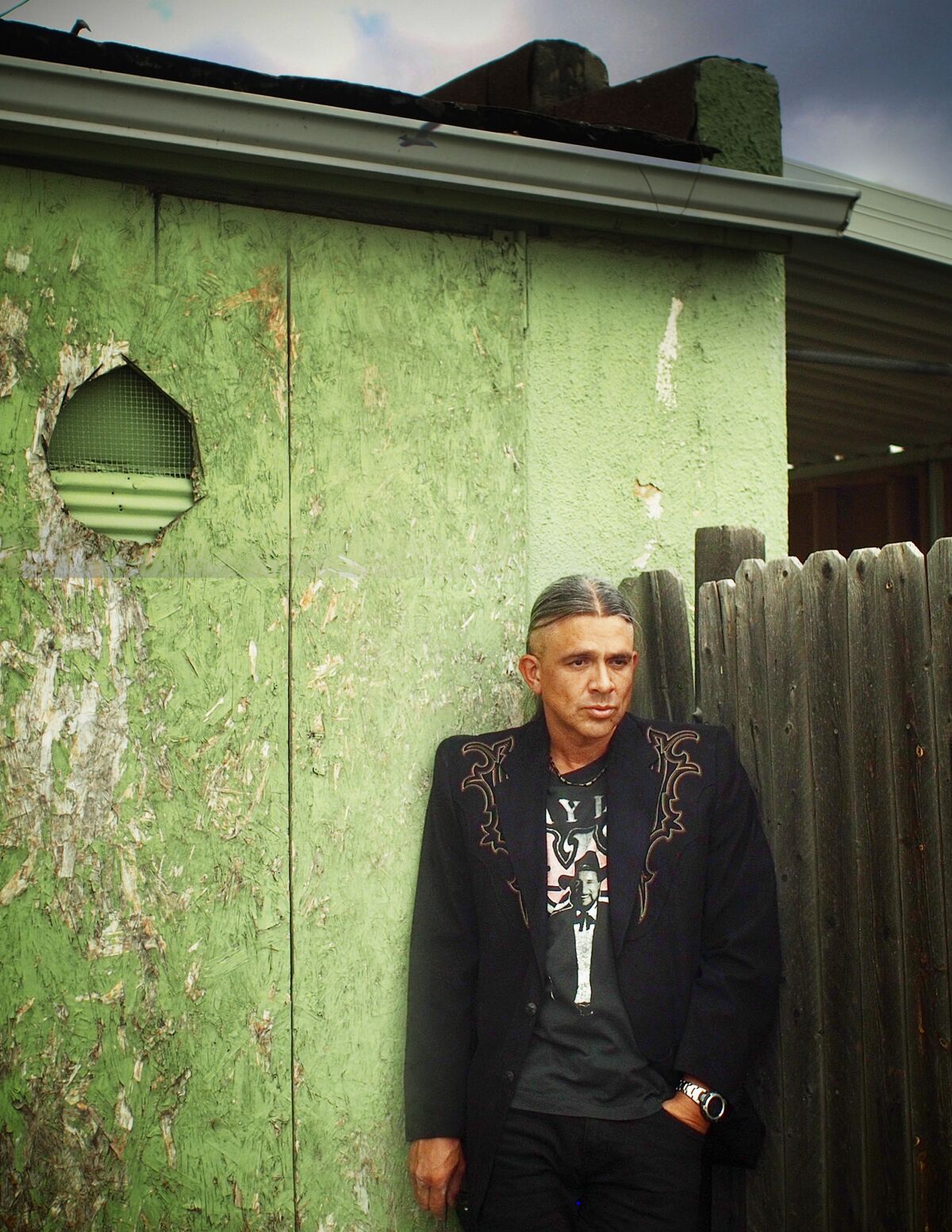 A man in an embellished jacket stands against a fence and an old building