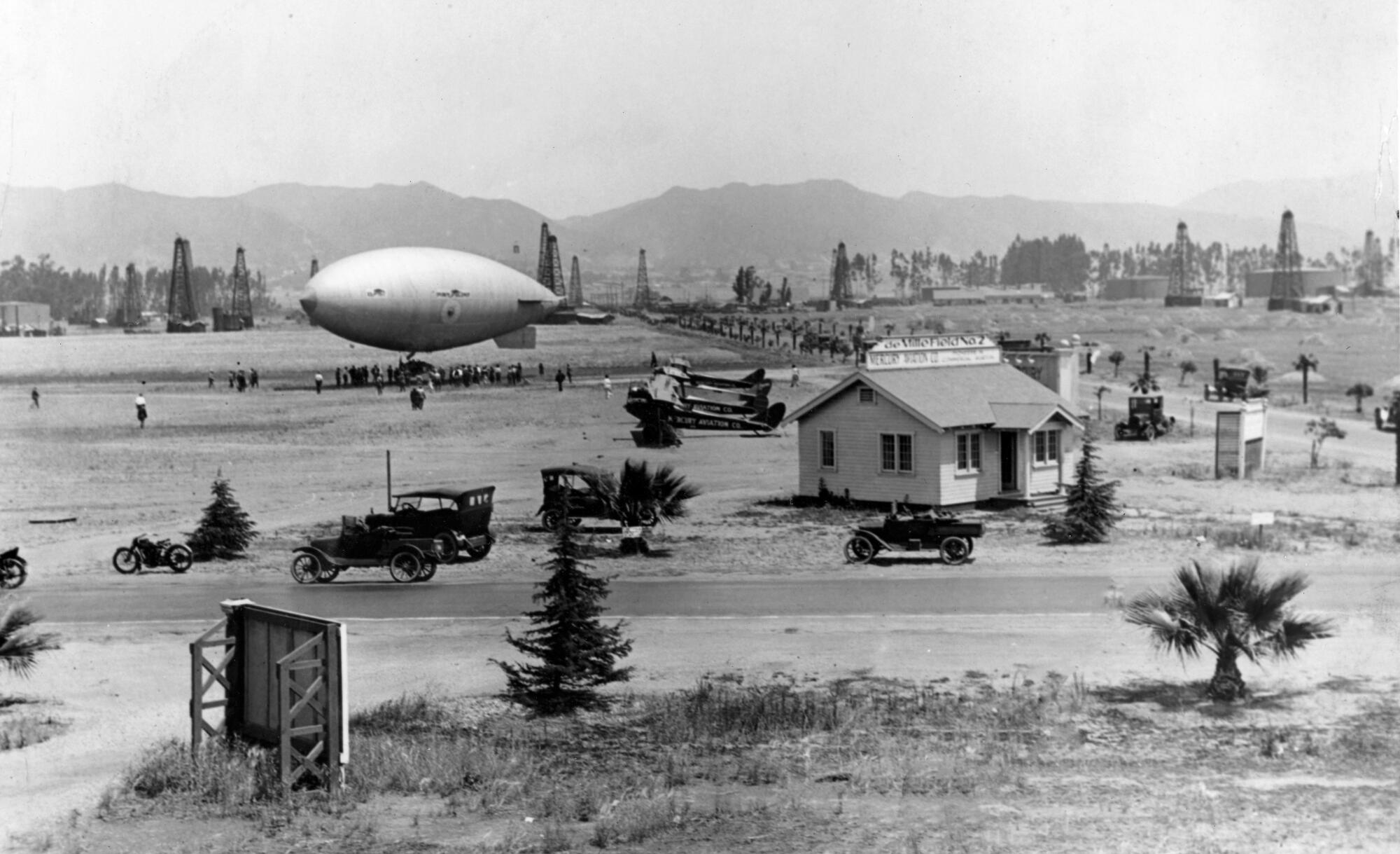 A vintage black and white photo shows a zeppelin in an airfield with oil derricks and mountains in the background