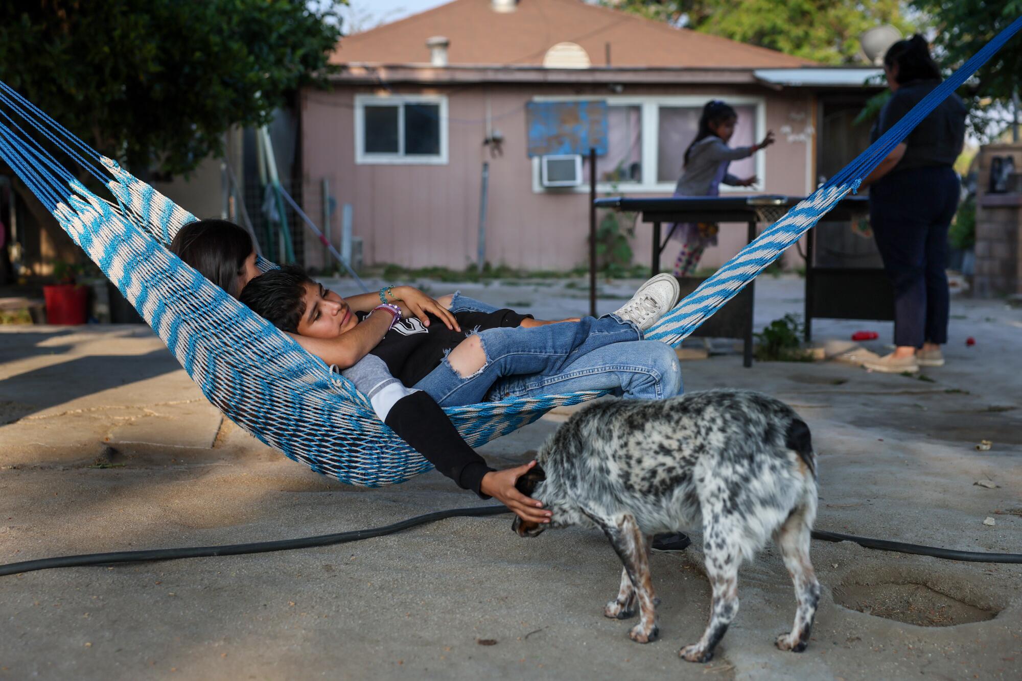 Two children swing in a hammock while petting their dog. 