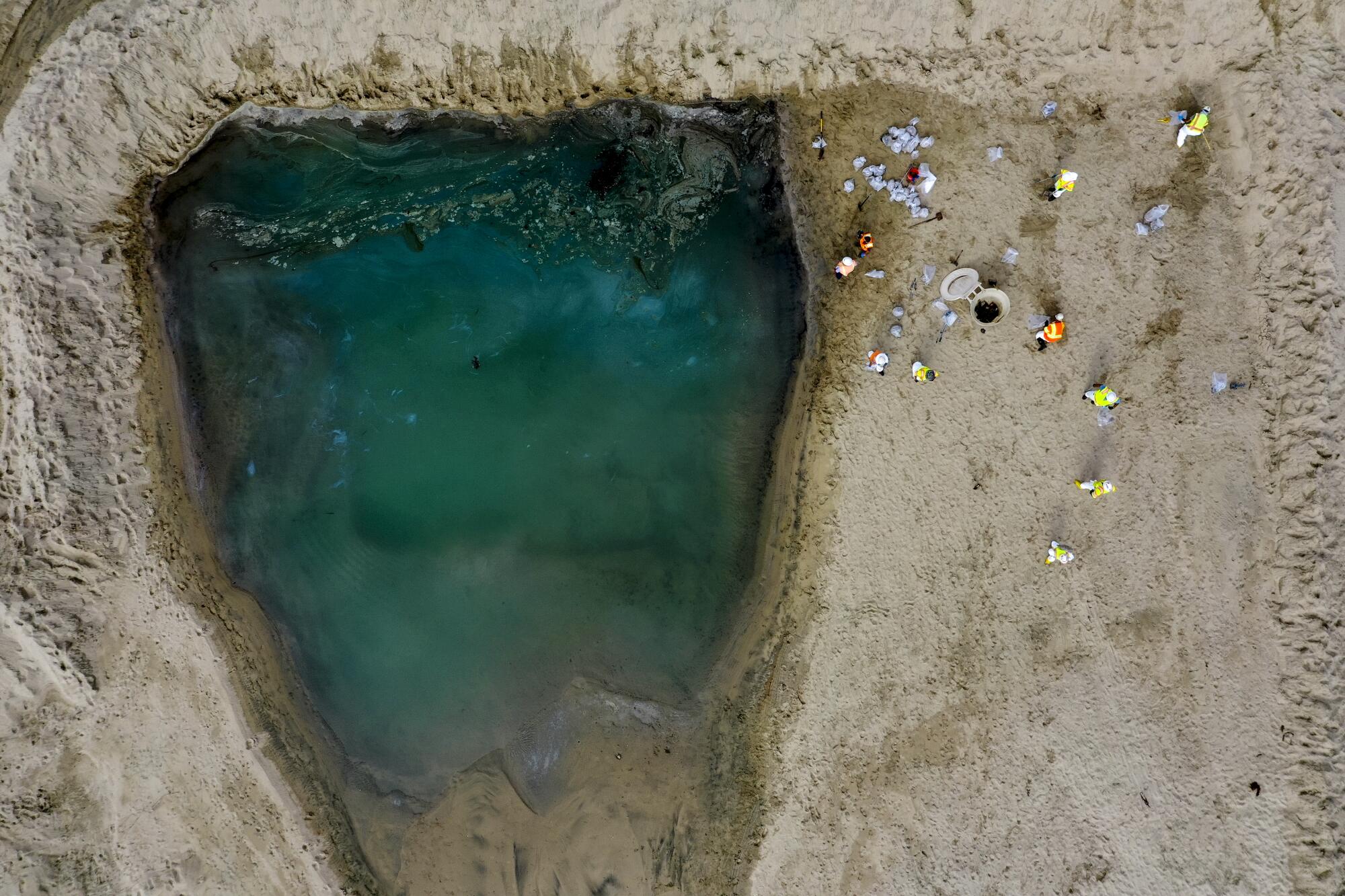  An aerial view of people gathered near the edge of a blue-green body of water surrounded by sand 