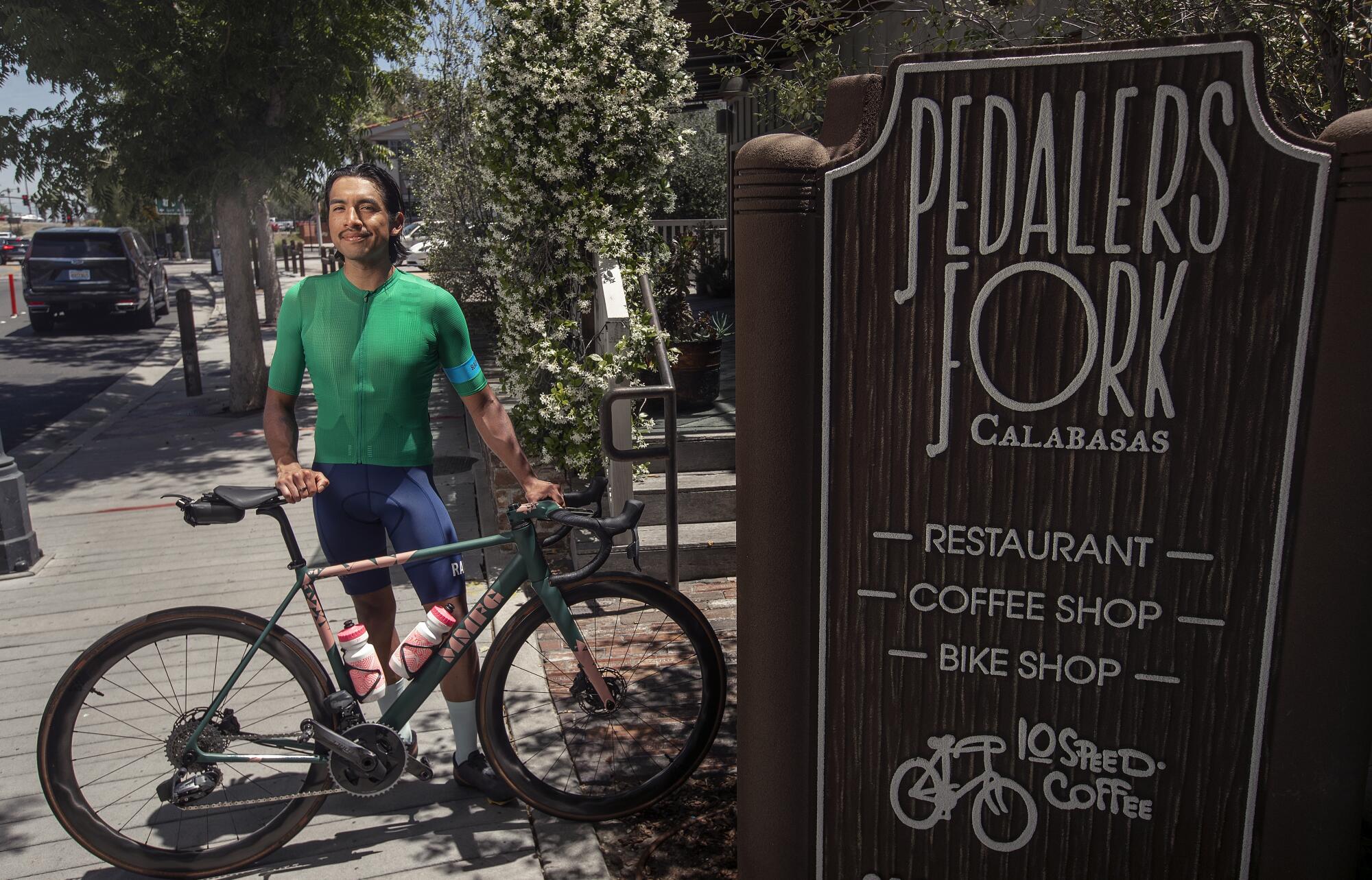 A man stands with a bicycle in front of a restaurant sign that reads "Pedalers Fork."