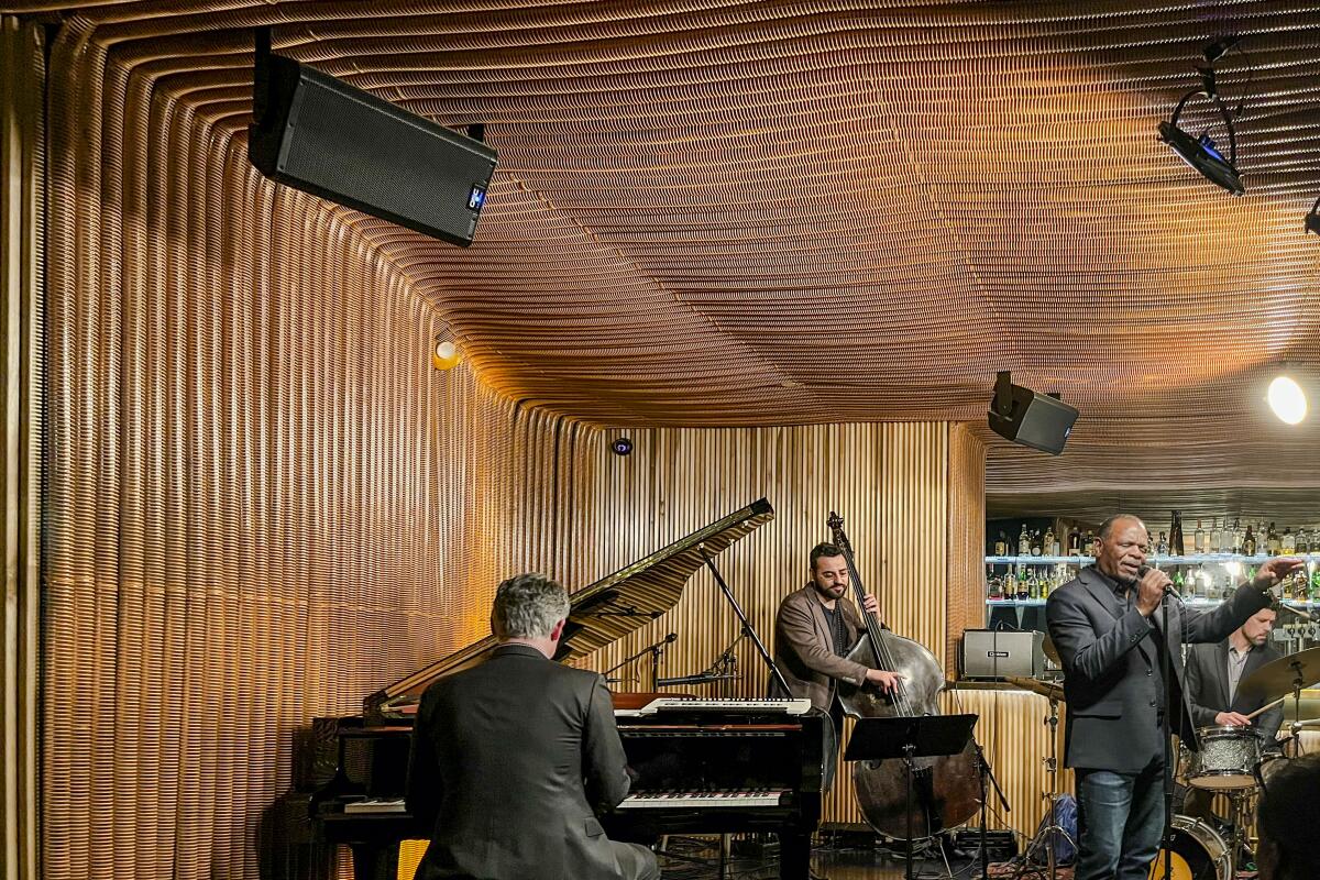 A man sings while two other men play an upright bass and a piano.
