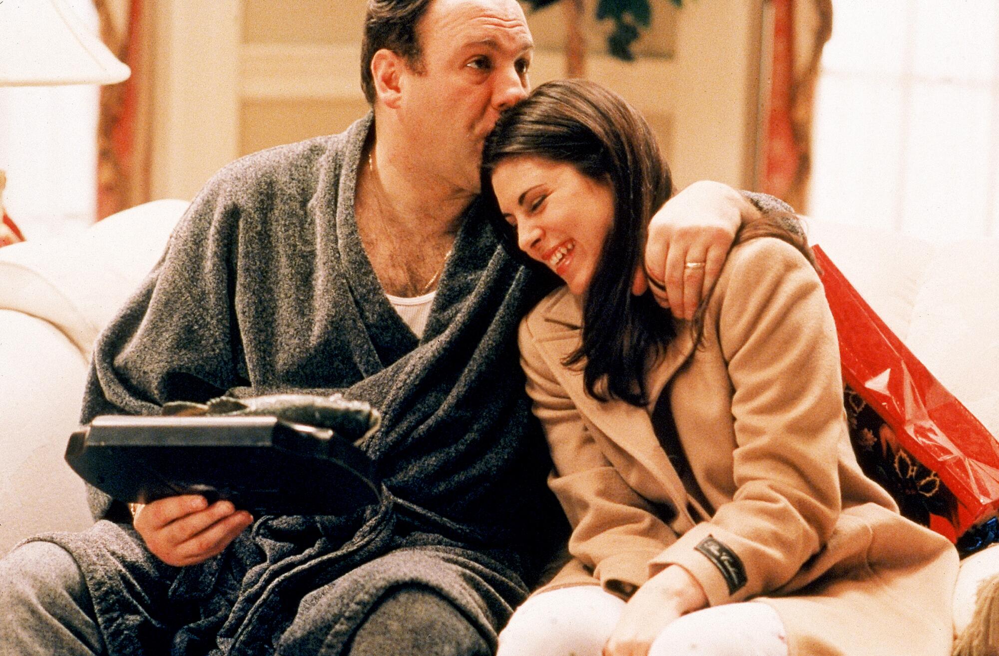 A man in a bathrobe holds a gift from his daughter and kisses her on the head. They are seated side by side on a couch.