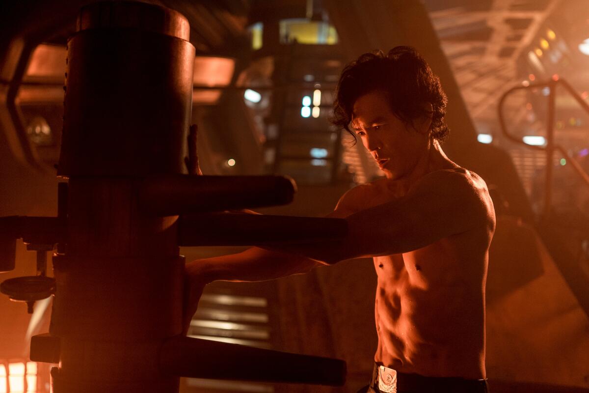 A shirtless, muscular John Cho performs a repair on a machine.