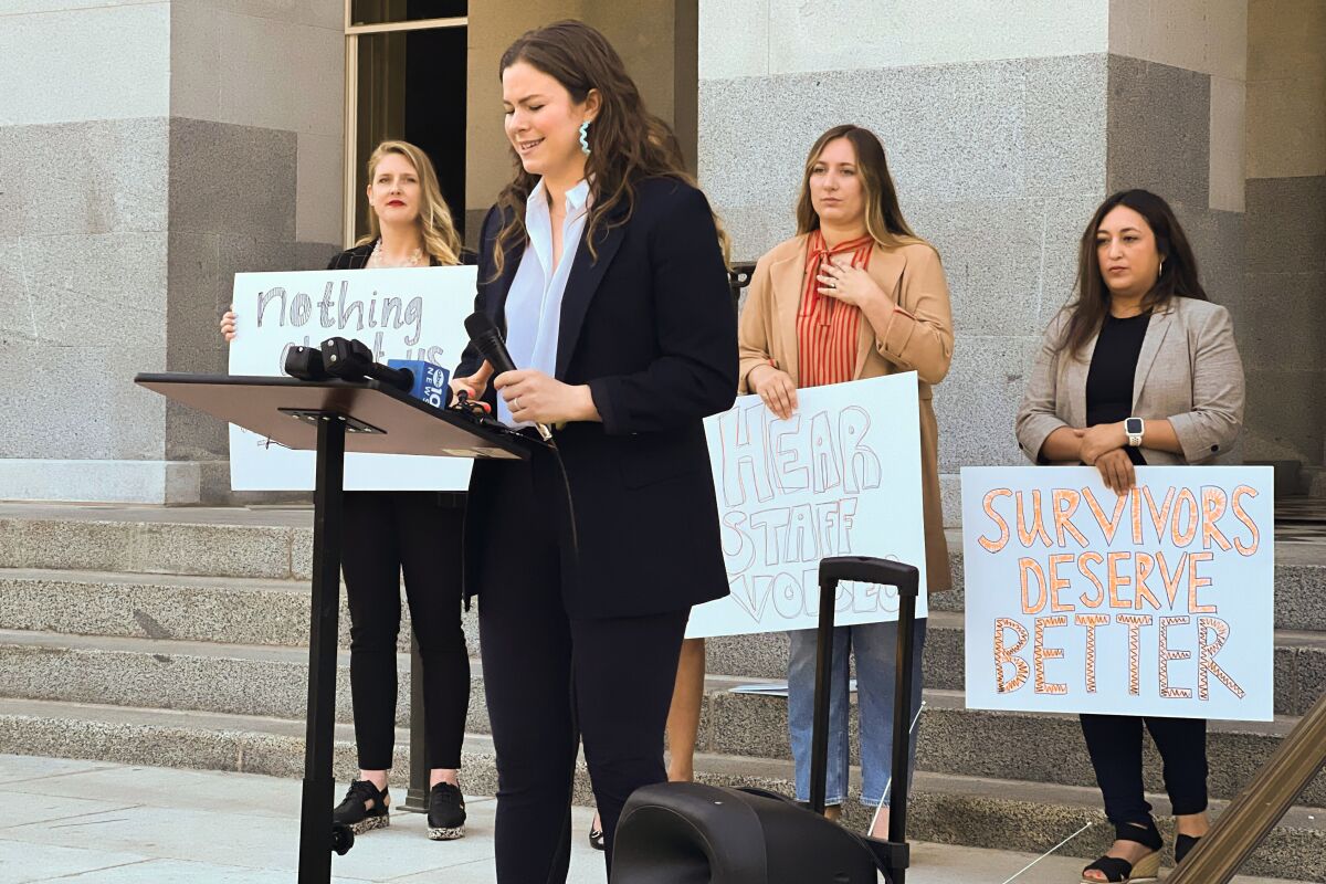 A woman stands at a lectern while three women holding signs stand behind her.