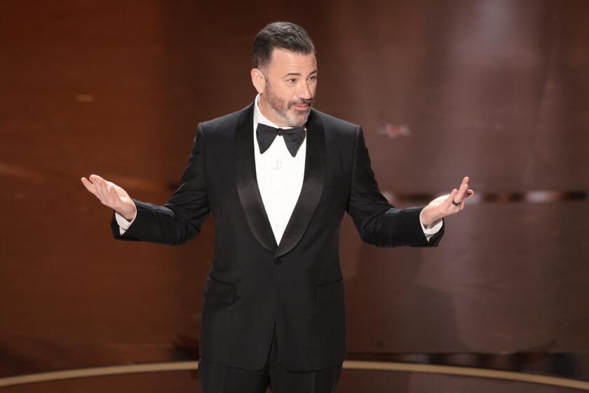 Jimmy Kimmel in a black tuxedo with a bowtie, standing on a stage with both of his hands out