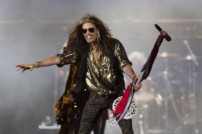Un hombre de cabello largo con atuendo de estrella de rock actúa en el escenario mientras sostiene un pie de micrófono en su mano izquierda