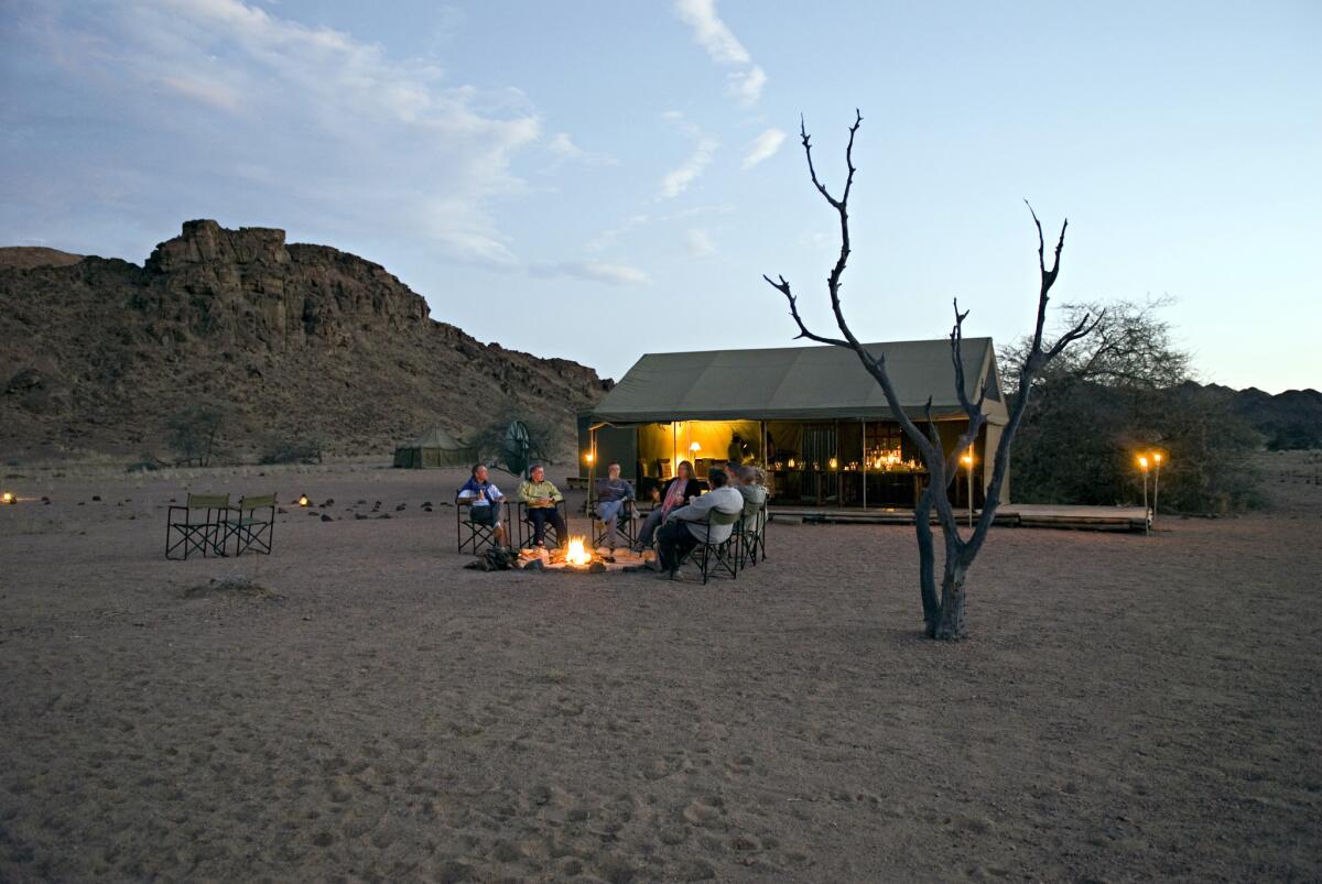 Visitors and guide sit around fire in folding camp chairs at sunset at Hoanib Camp in Kaokoland, Namibia.