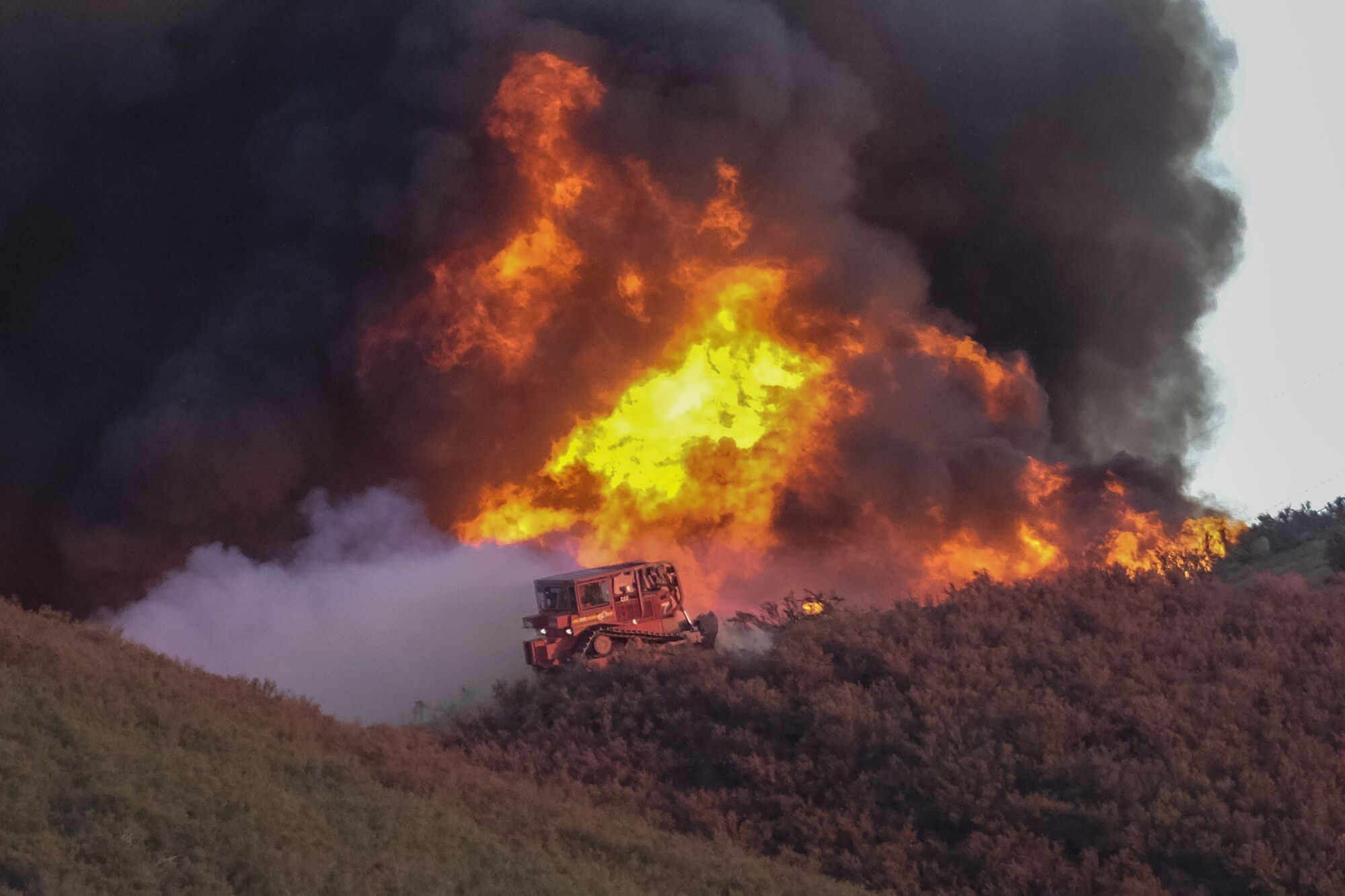 A bulldozer works to build a fire line while flames and smoke rise behind it.