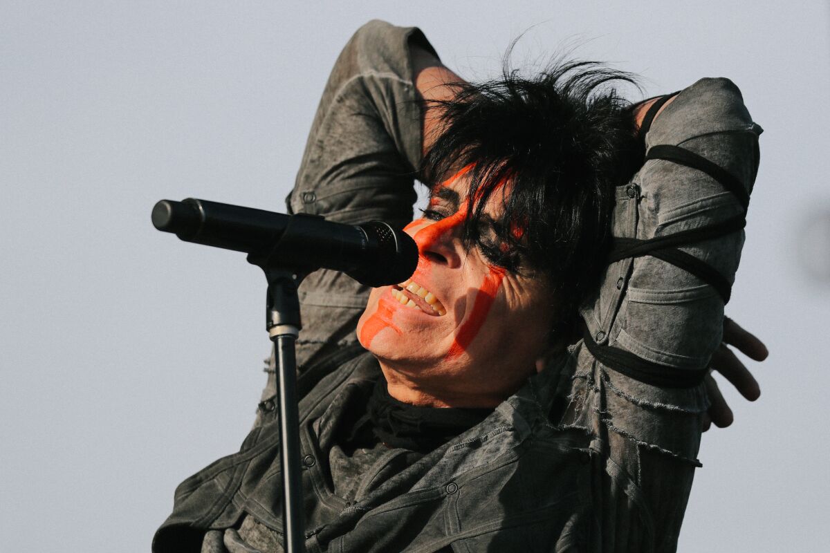 A man at a microphone with dyed black hair and streaked orange face paint stands with his arms behind his head.