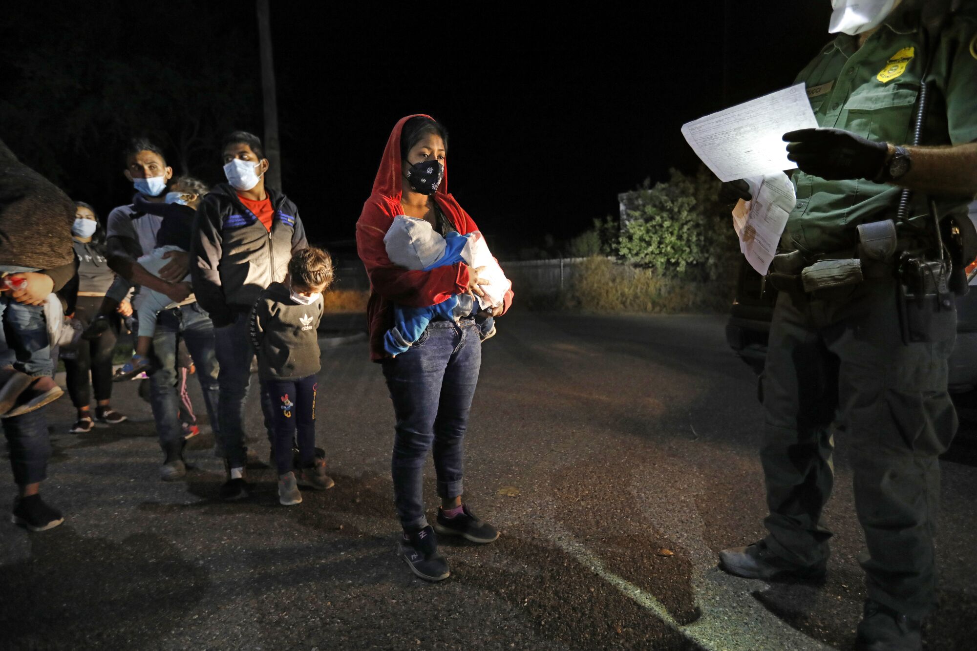 A line of migrant adults and children standing before a Border Patrol agent at night.