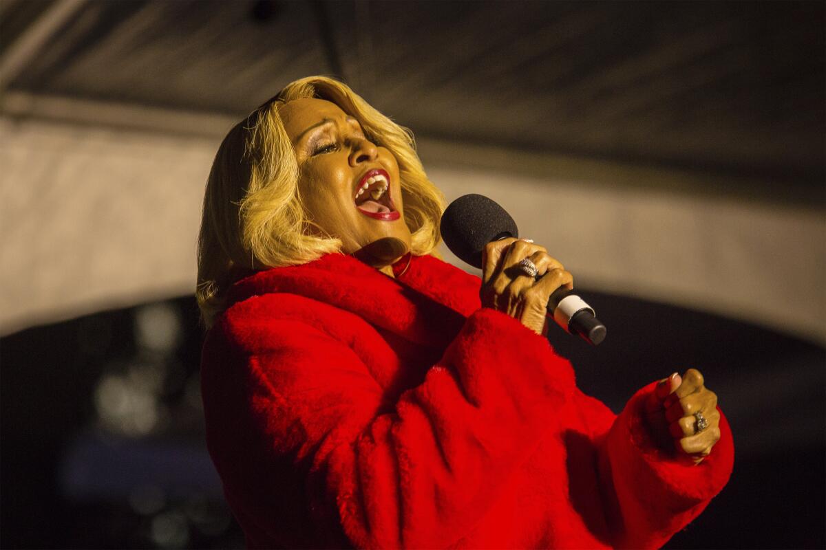 An older woman in a furry red coat sings into a microphone at a holiday event.