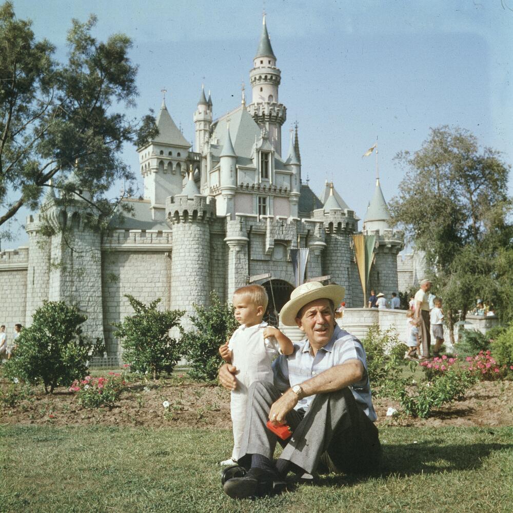 Walt Disney sits on a grassy lawn with his grandson, in front of the castle