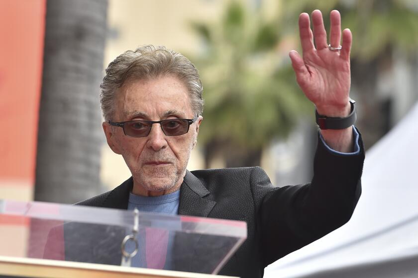 Frankie Valli in sunglasses, a blue shirt and a dark suit jacket holding up his left hand as he approaches a podium.