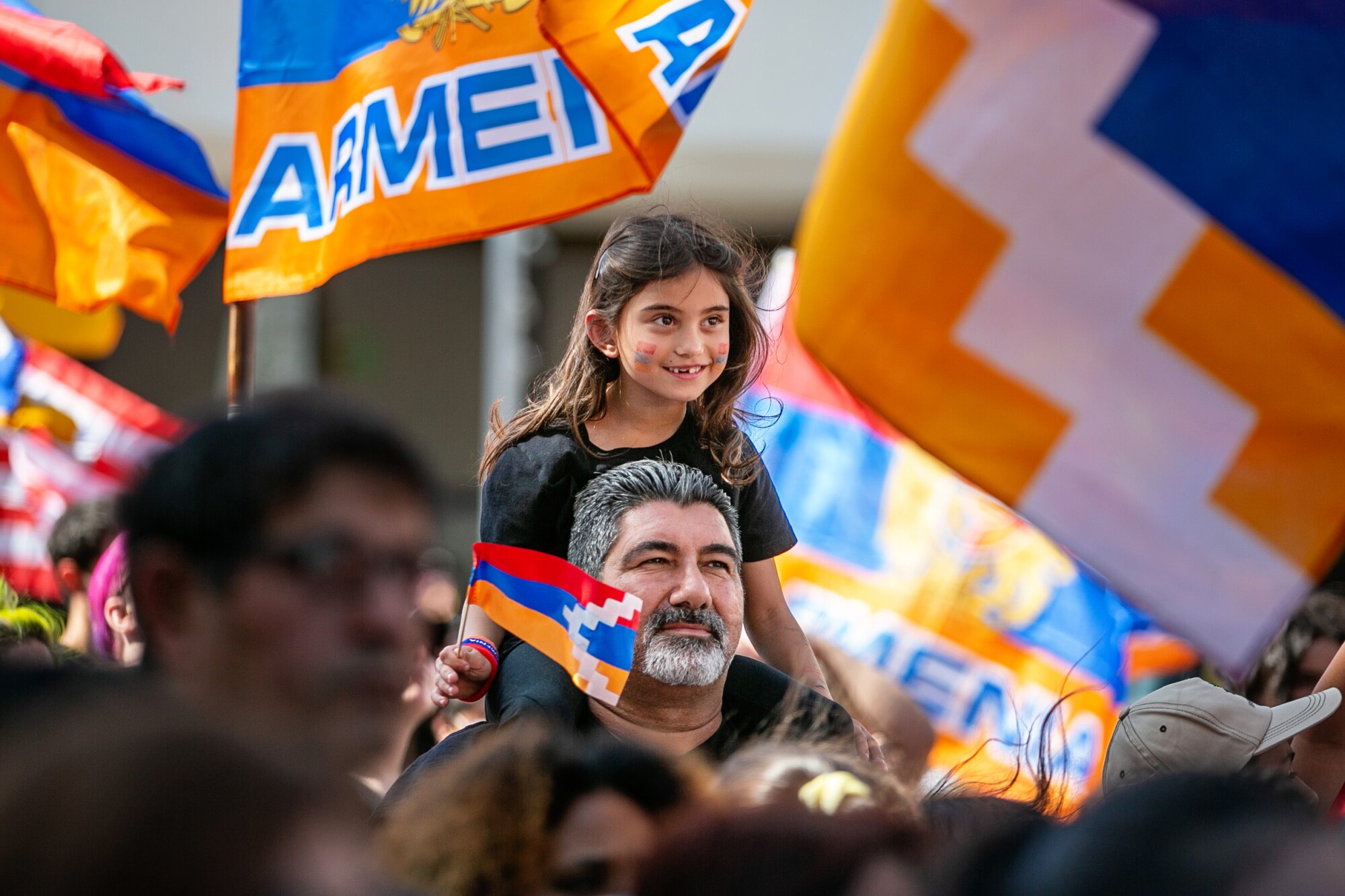 A girl sits on a mans shoulders as protesters hold Armenian flags