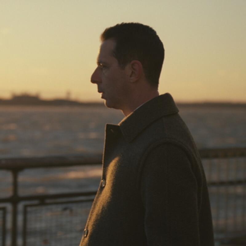Jeremy Strong (in character as Kendall Roy) looks out over a body of water while wearing a winter coat.