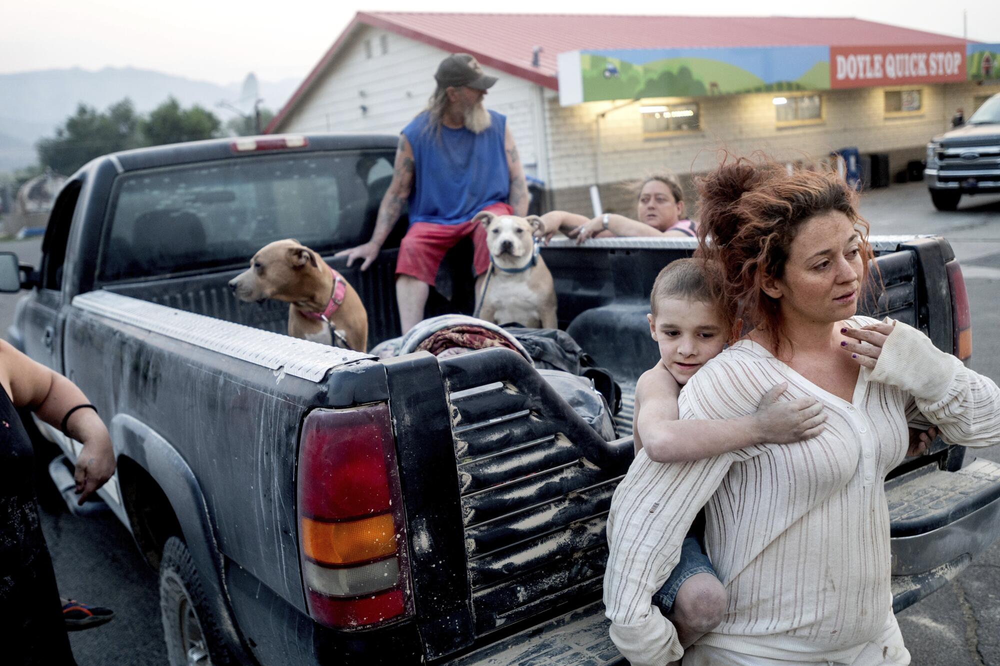 A woman holds a child as an older man and two dogs sit in the bed of a pickup truck.