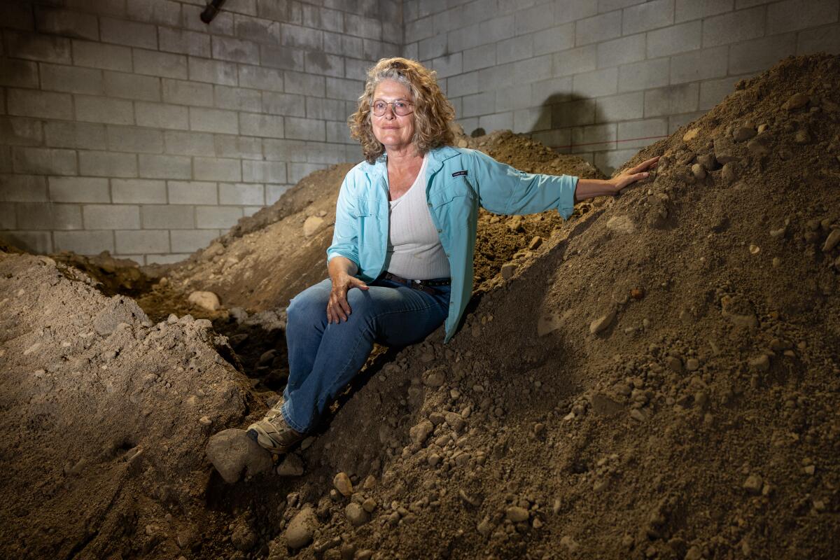 Artist Lauren Bon, who has blonde curly hair, sits on a mound of earth inside a warehouse building.