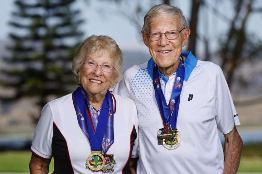 An older couple wearing pickleball uniforms, wearing award medals and holding rackets