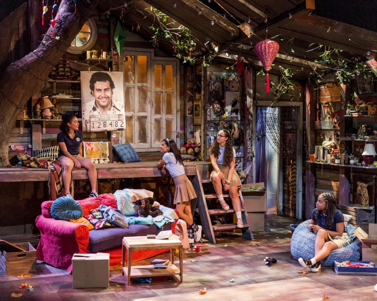 Girls are sprawled around a stage set meant to look like a woody clubhouse with a photo of Pablo Escobar at center