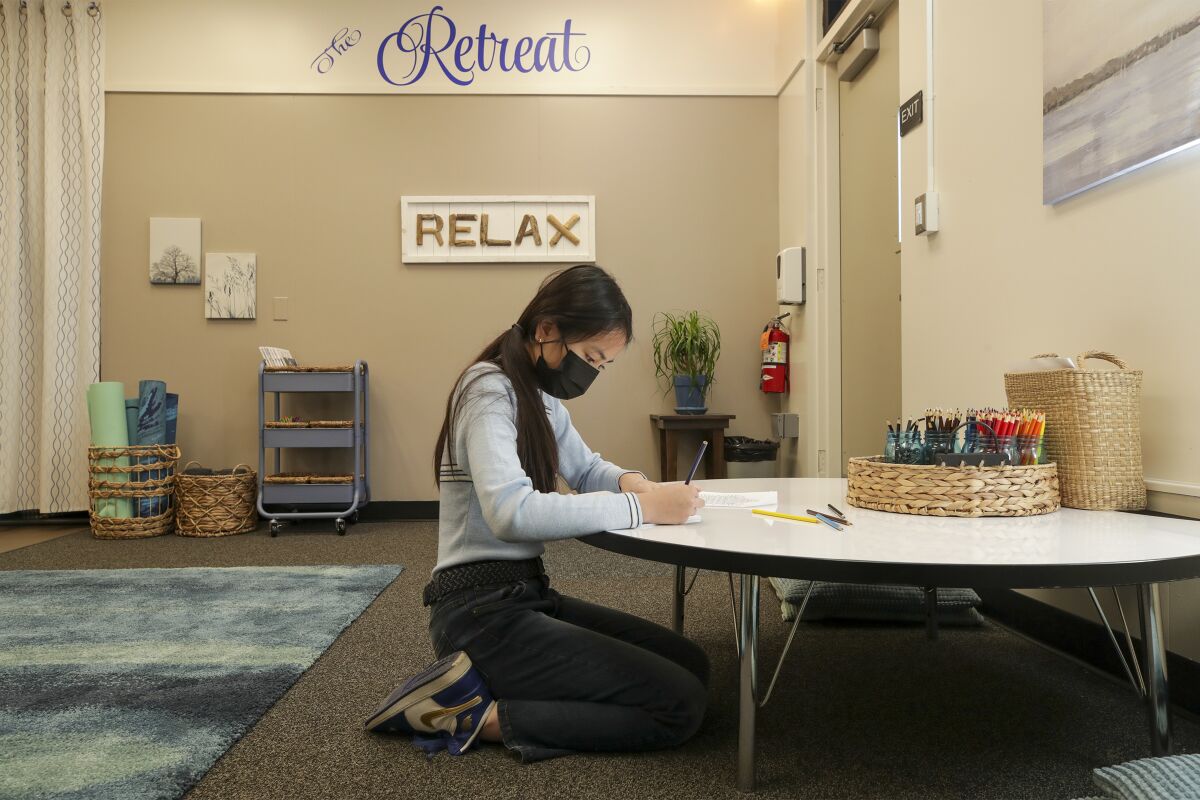 A masked student sits at a low table and uses colored pencils in the calming room at her school.