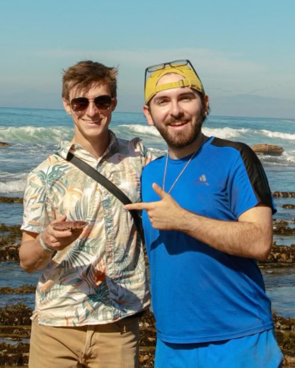 Two young men pose for a photo with the ocean behind them.