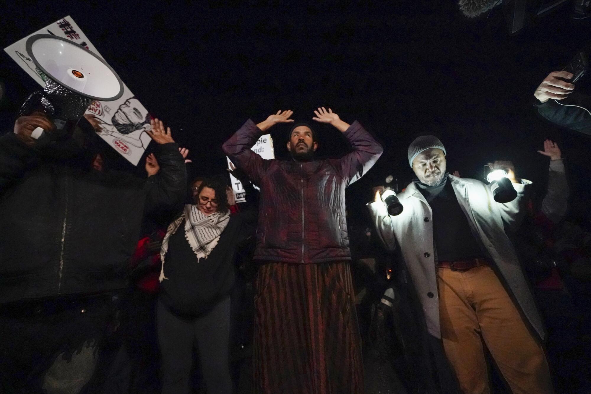 Several people protesting in the dark, some with their hands up 