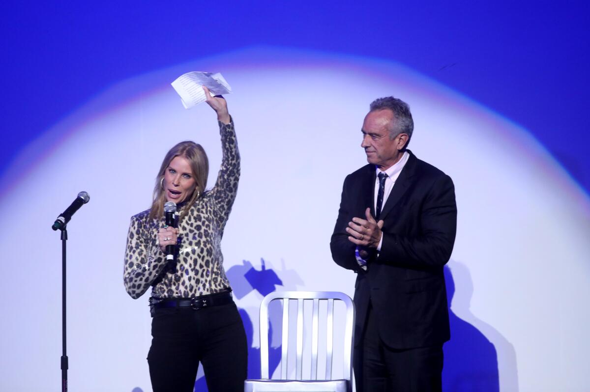 Cheryl Hines holds a piece of paper high as she speaks into a mic, sharing a spotlight with a clapping Robert F. Kennedy Jr.
