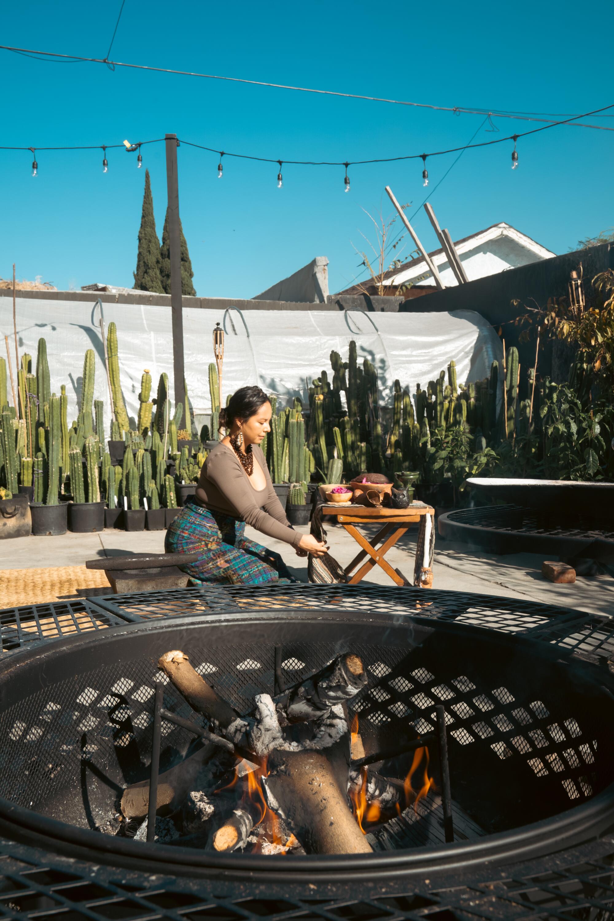 A woman kneels near a table while a fire pit burns
