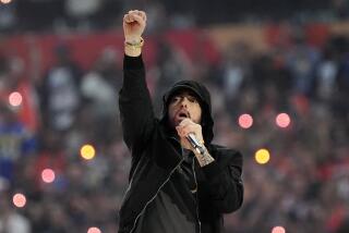 Eminem wearing a black hoodie with his right arm and hand in a fist performing on stage