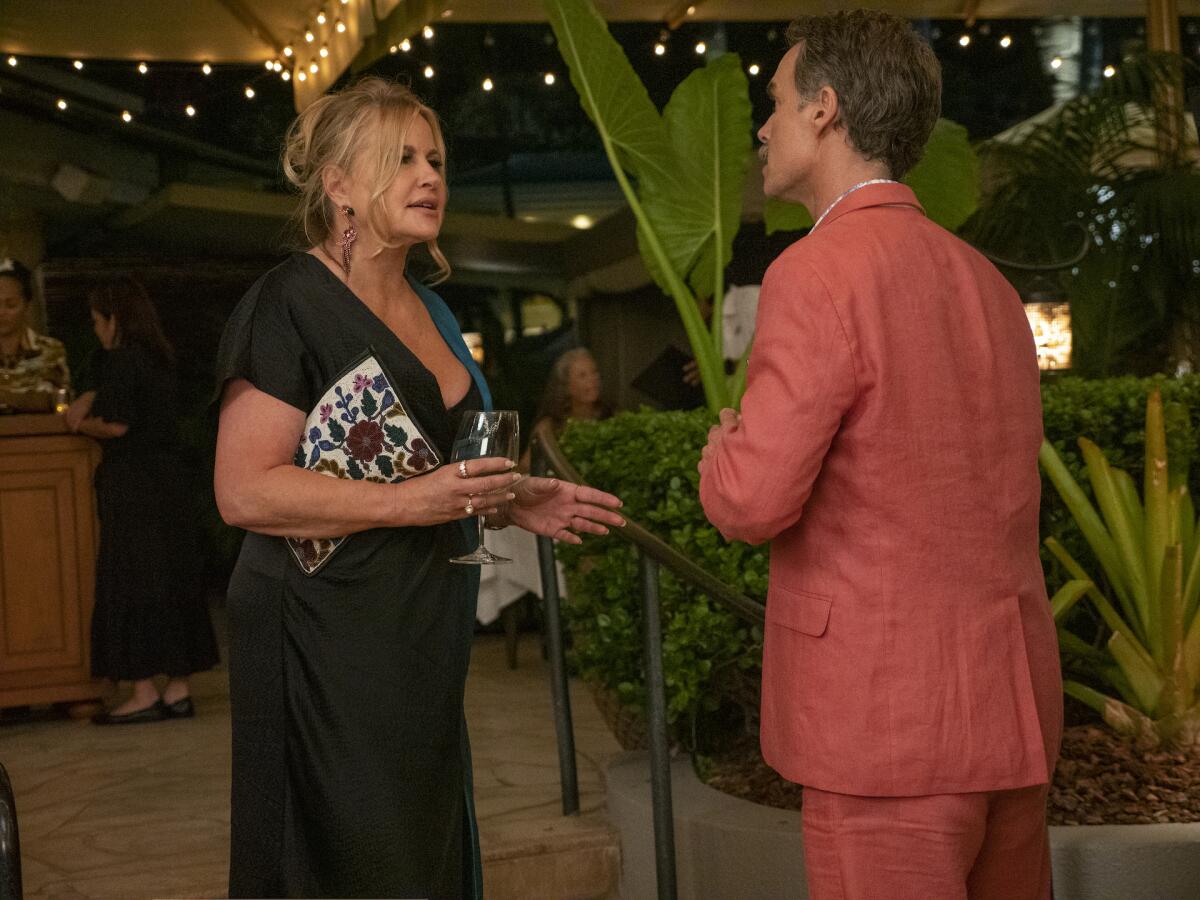 A woman in a black dress talks to a man in a coral-colored suit at a tropical resort.