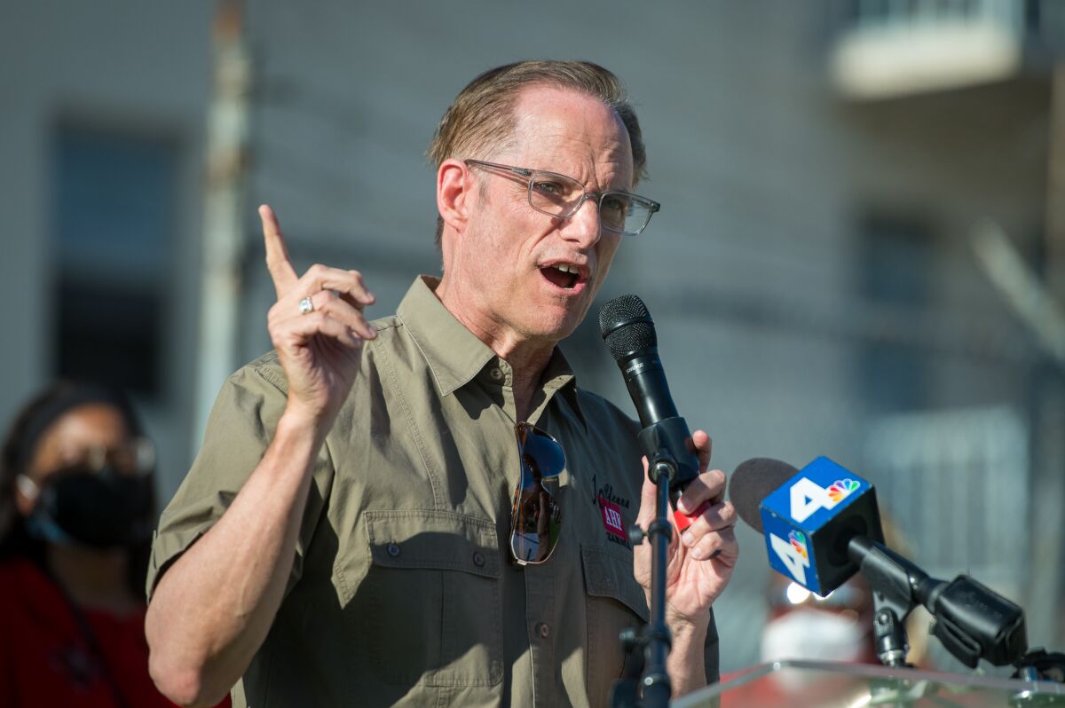 A man raises his right hand, index finger extended, as he speaks into a microphone.