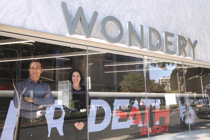 WEST HOLLYWOOD, CA - OCTOBER 27, 2020 - Wondery CEO Hernan Lopez, left, and COO Jen Sargent stand in the display window for the company as podcasts of their programming projects on an LED screen, background, in West Hollywood on October 27, 2020. Lopez is the founder and CEO of Wondery, creates and curates immersive storytelling-based podcasts including Dirty John, Sword & Scale, Hollywood & Crime and Dr. Death, season two. (Genaro Molina / Los Angeles Times)