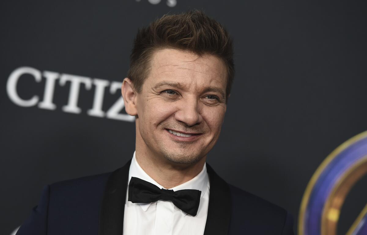 Jeremy Renner smiling in a black suit and bowtie against a black background