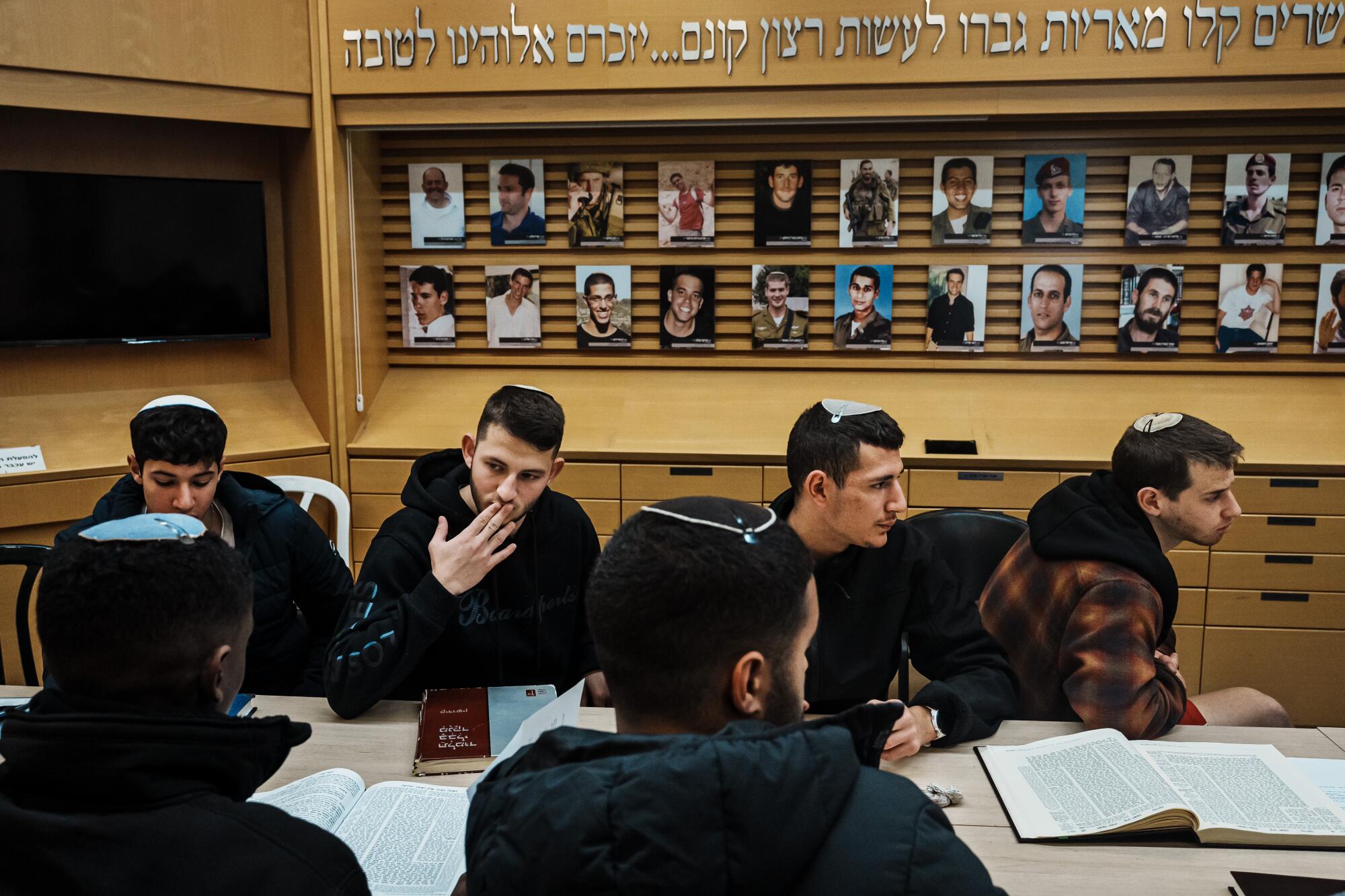 Young men in yarmulkes sitting at a table with books and papers, in front of a display with two rows of portraits