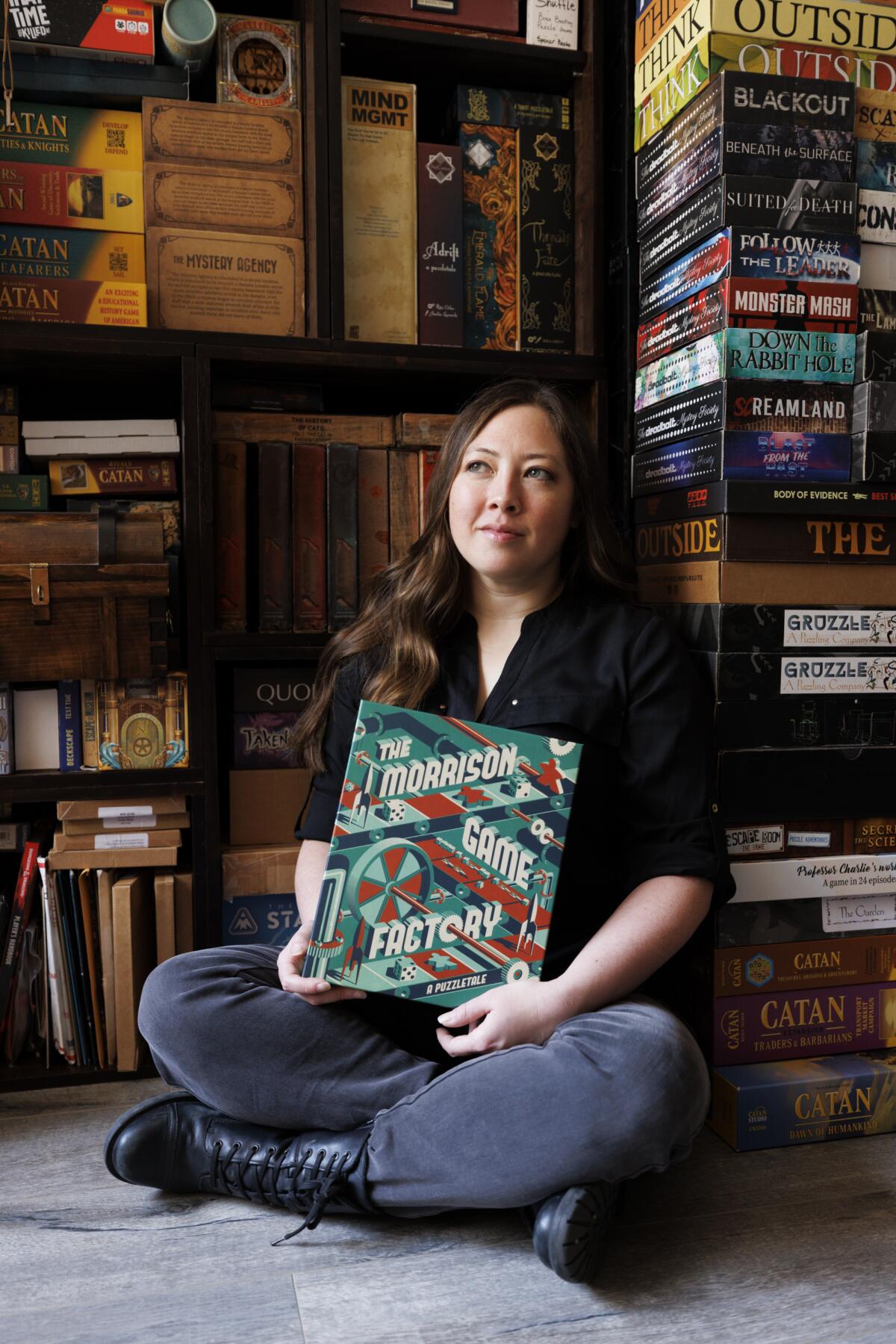 Lauren Bello sits cross-legged on the floor, holding her game, “The Morrison Game Factory,” in its box