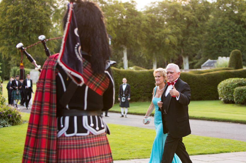 A man and woman in formalwear walk past a kilt-clad bagpiper.