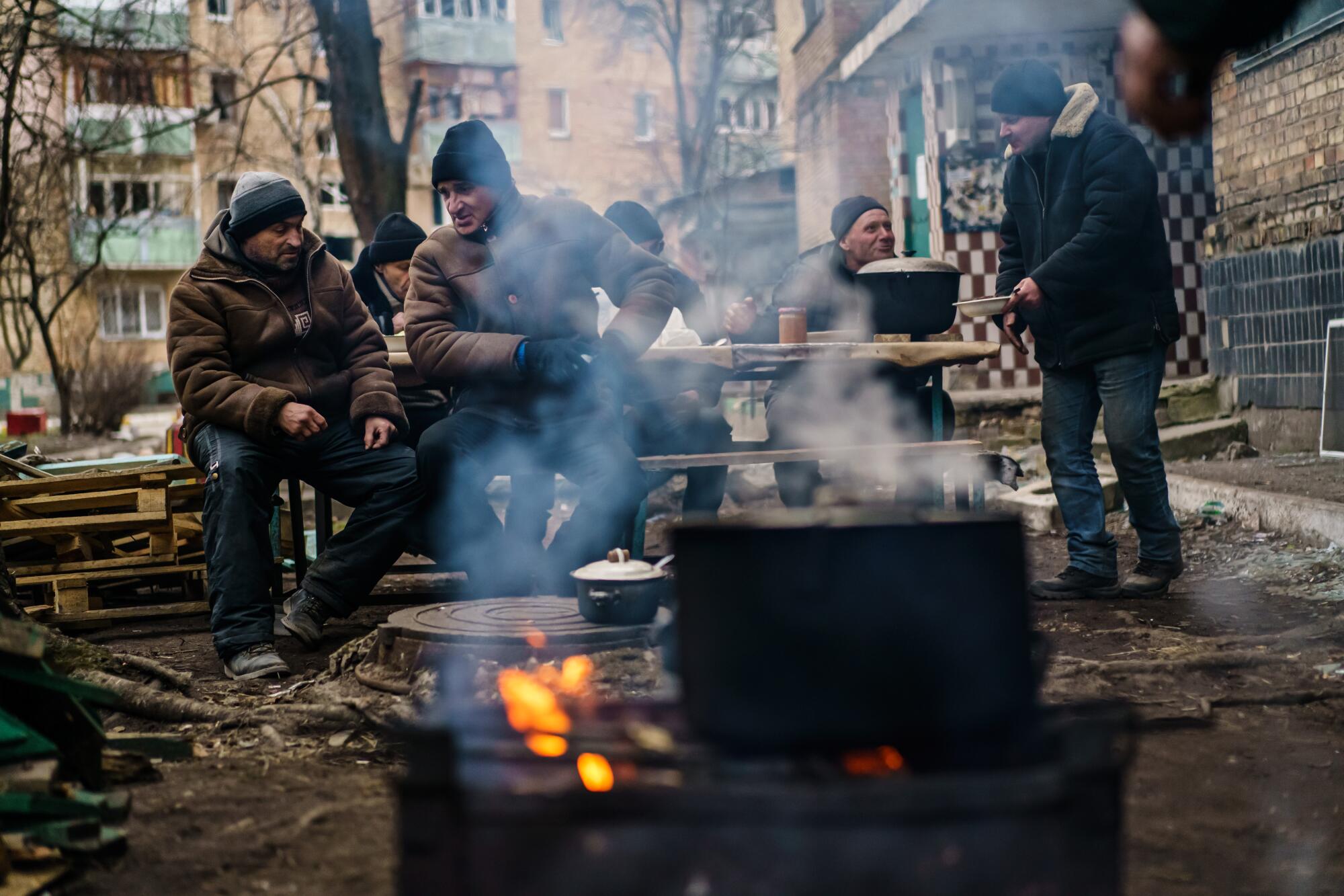 Men in coats and hats gather around outdoor fires with cooking pots