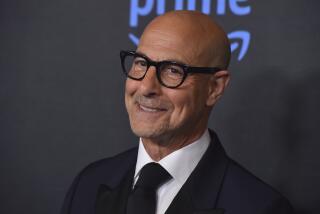 Stanley Tucci smiles while wearing glasses and a black suit and tie.