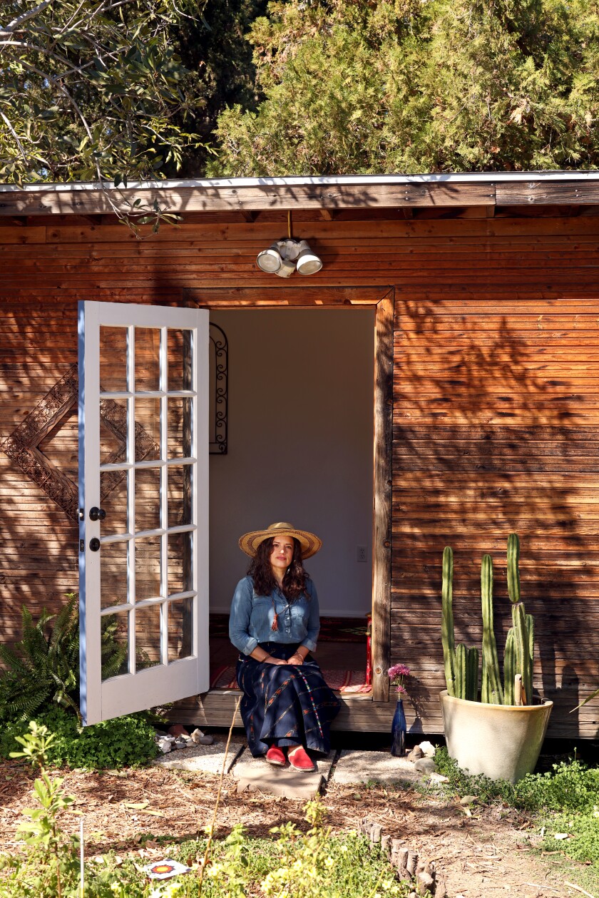 A woman wearing a hat smiles while sitting in the doorway of a wood structure.