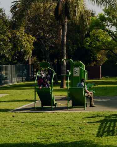 People sit on benches and play on playground equipment in a small park on a sunny day
