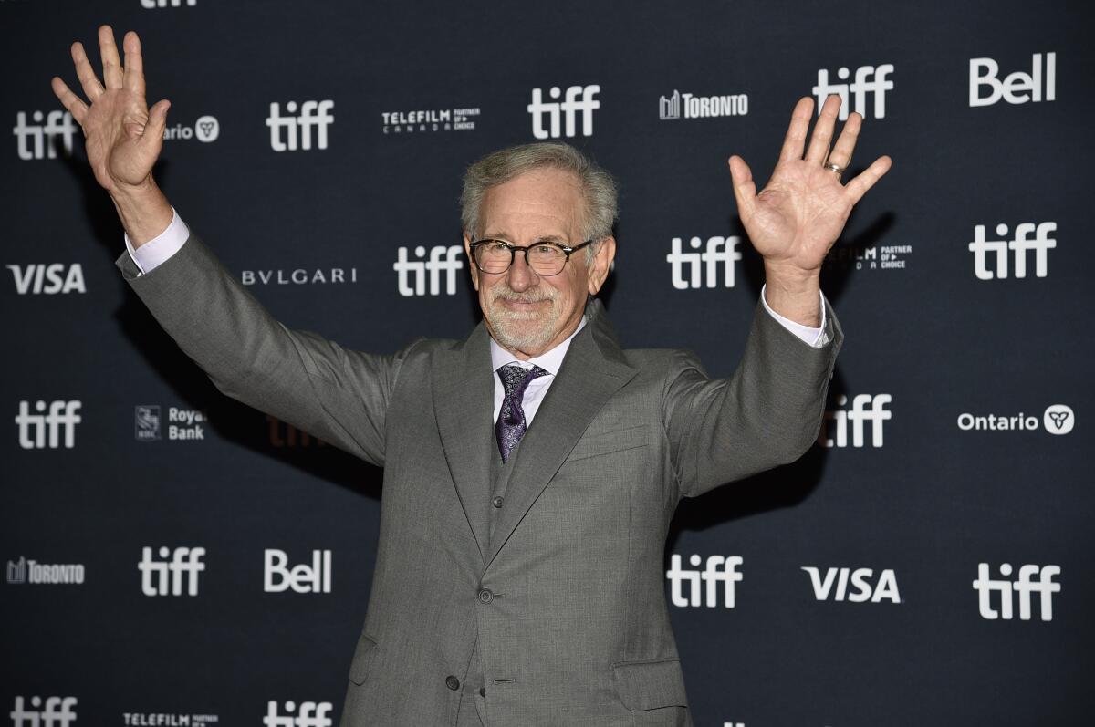 A man in a suit and round glasses waves 