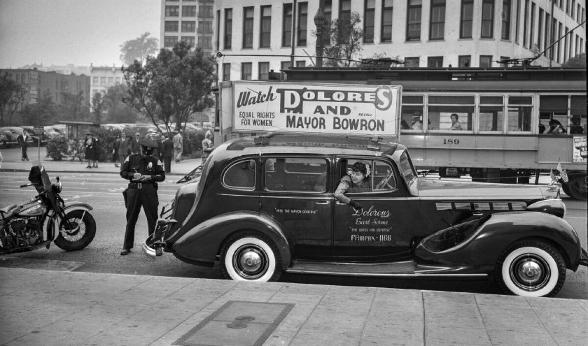 A woman sits in a vintage automobile topped with a campaign sign as a motorcycle officer writes a citation