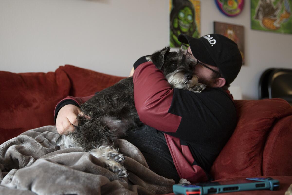 A man in a baseball cap cuddles a dog on a couch 