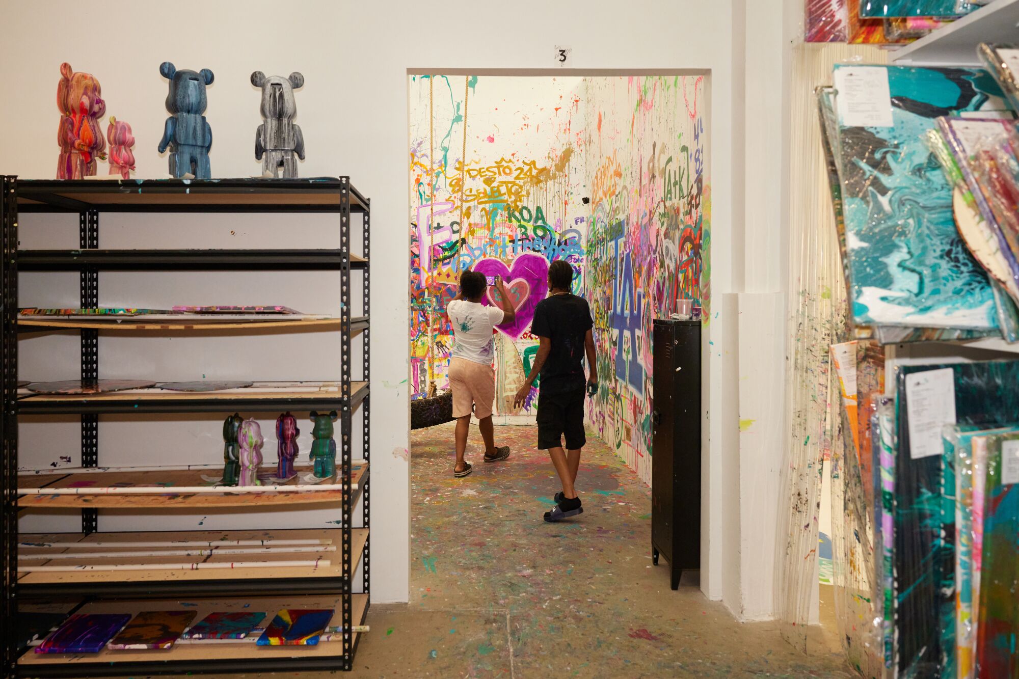 Two figures stand behind the doorway of an art studio with multicolor paint on the walls.